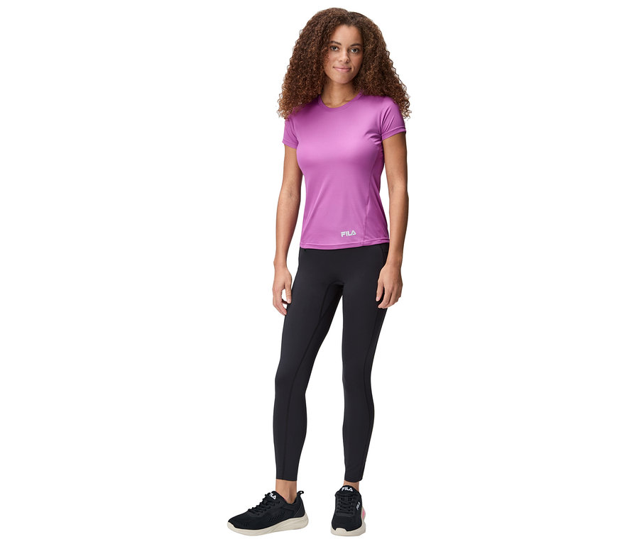 Žena stojí, má na sebe fialové tričko Fila, čierne legíny a čierne tenisky.
