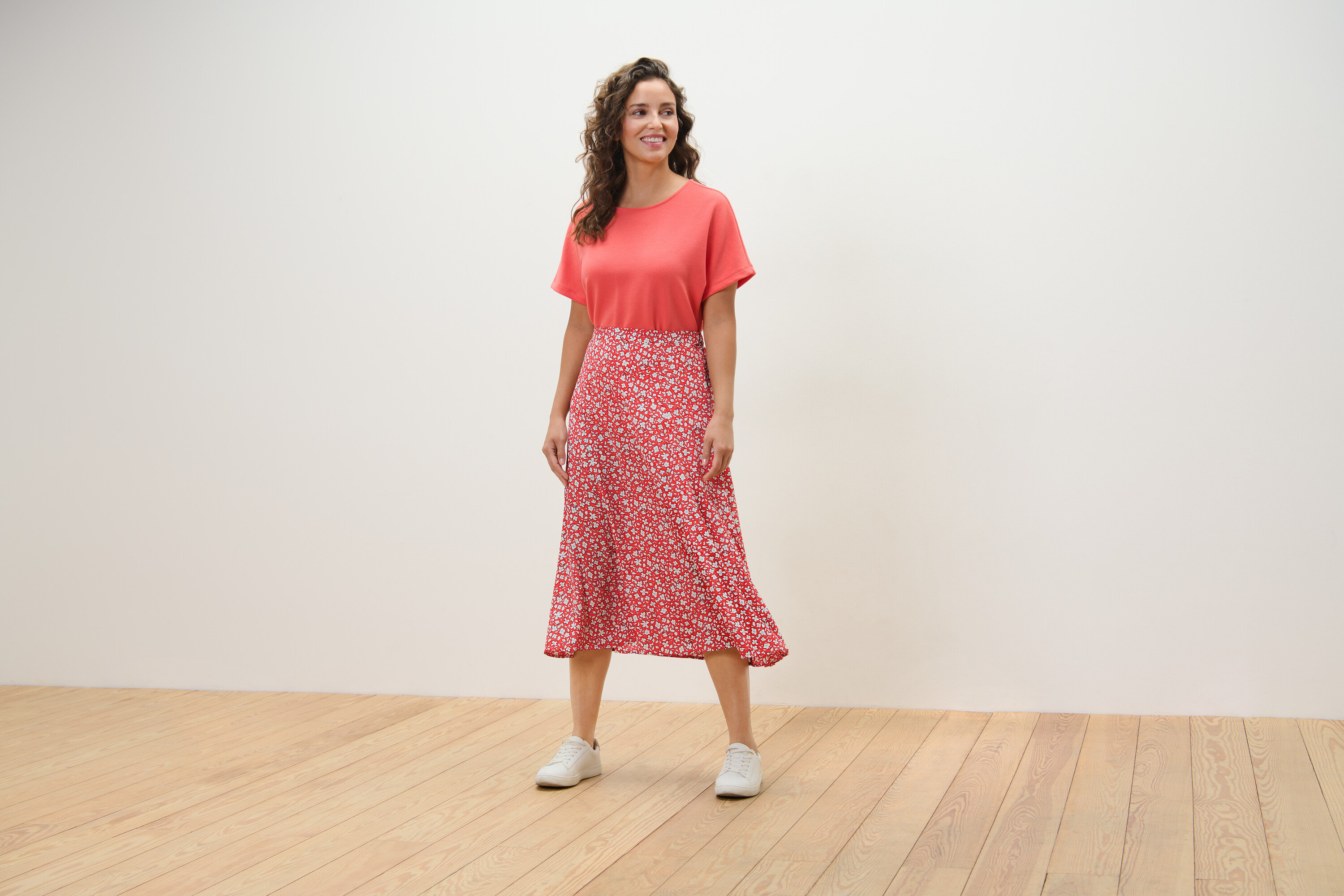 Žena stojí na drevenej podlahe a má na sebe koralovú sukňu s potlačou.