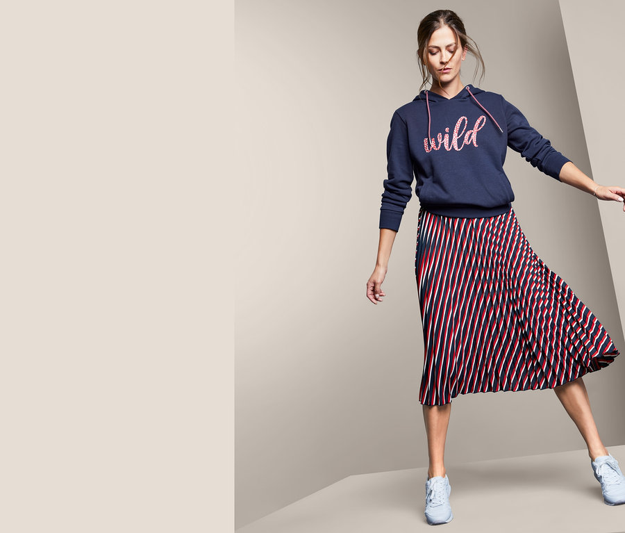Žena pózuje v plisovanej sukni a mikine s kapucňou s textovou potlačou.