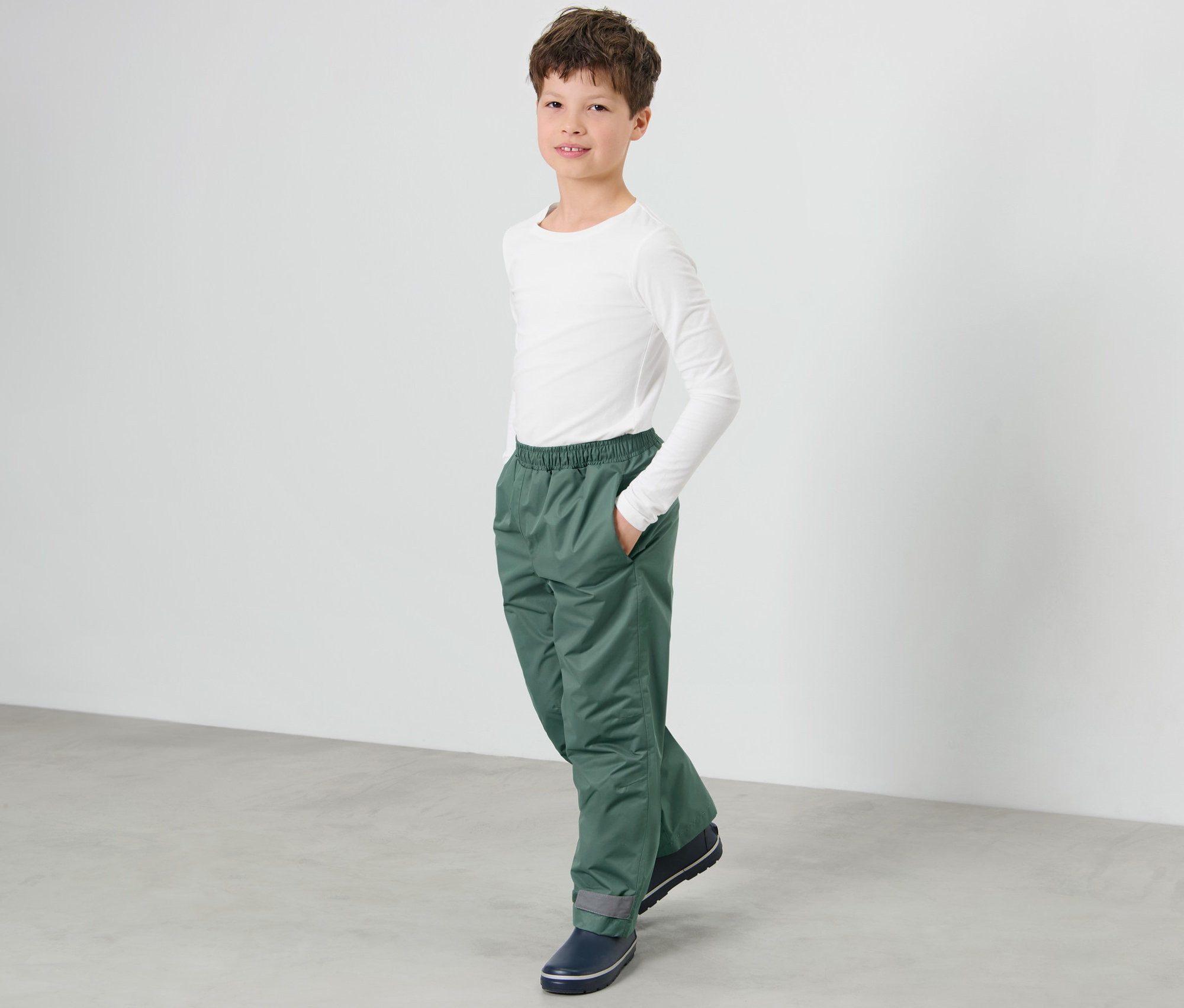 Chlapec stojí a má ruku vo vrecku detských nohavíc do dažďa.