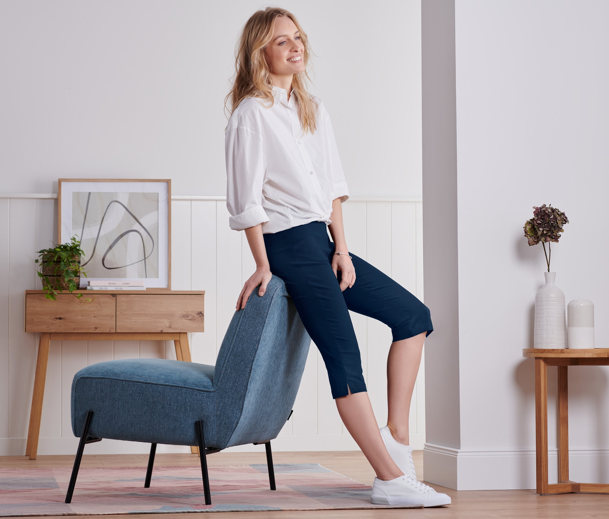 Žena sedí bokom na modrom kresle a má na sebe námornícke modré trojštvrťové nohavice.