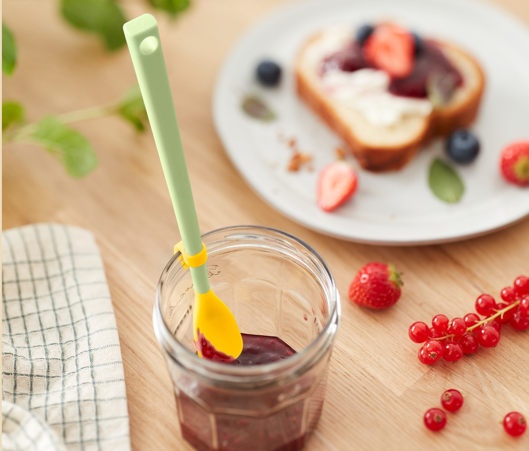 Džem v pohári so špeciálnou silikónovou lyžicou. V pozadí je toast s ovocím.