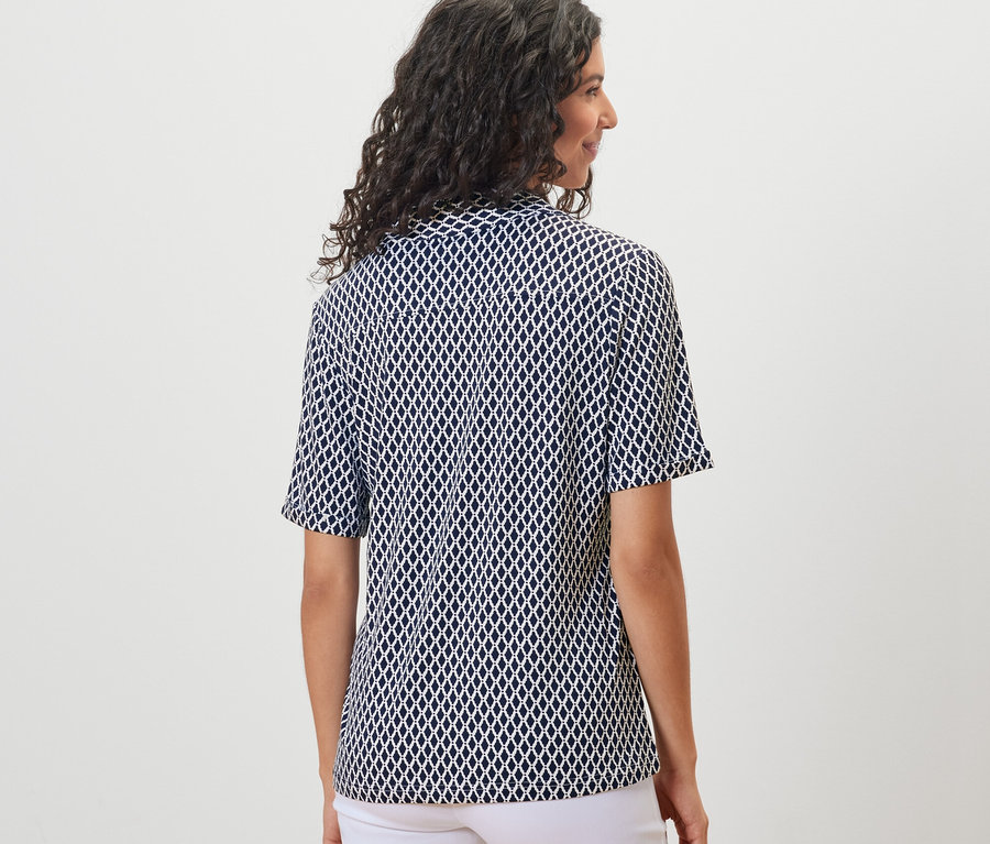 Pohľad zozadu na ženu s tmavými vlnitými vlasmi v blúzkovom tričku s potlačou s gombíkovou légou a bielych nohaviciach.