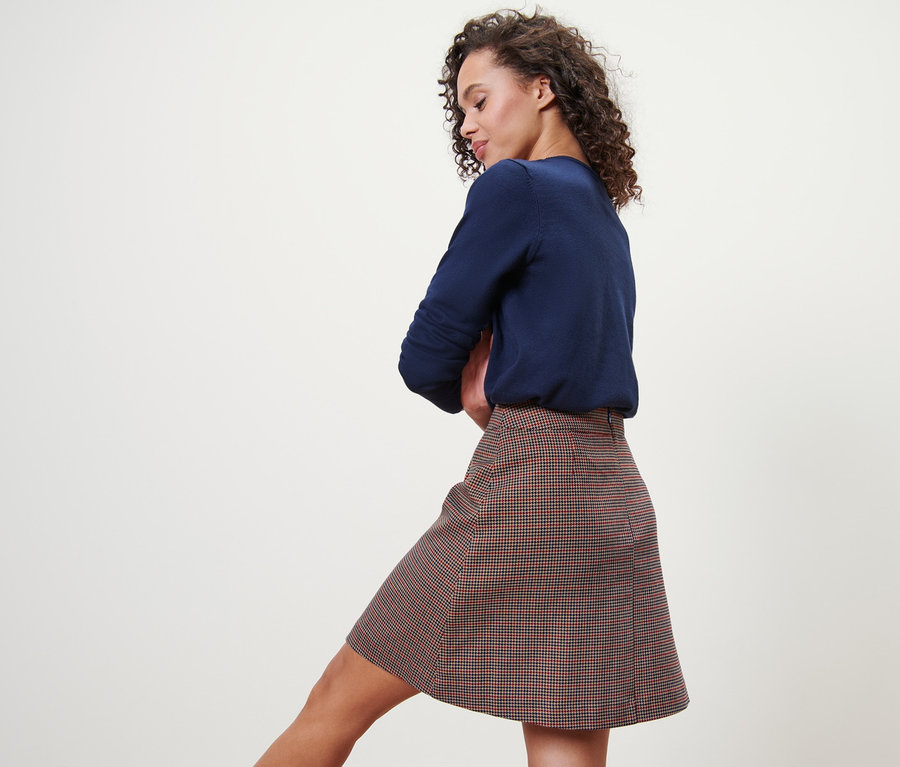 Žena pózuje v károvanej tkanej sukni a modrom svetri.