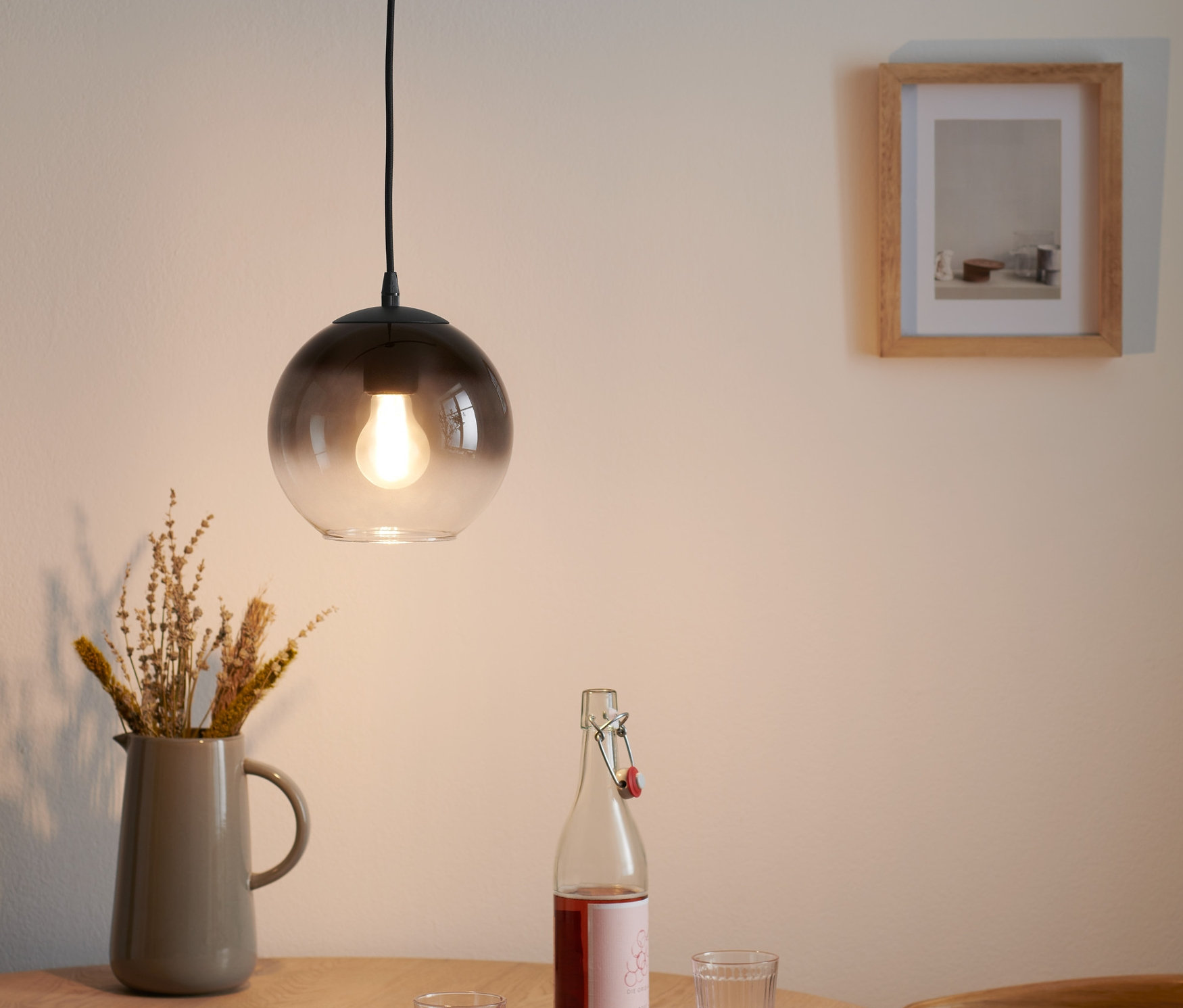 Závesné svietidlo s tienidlom z dymového skla visí nad dreveným stolom, na ktorom je váza s kvetmi, fľaša a dva poháre.