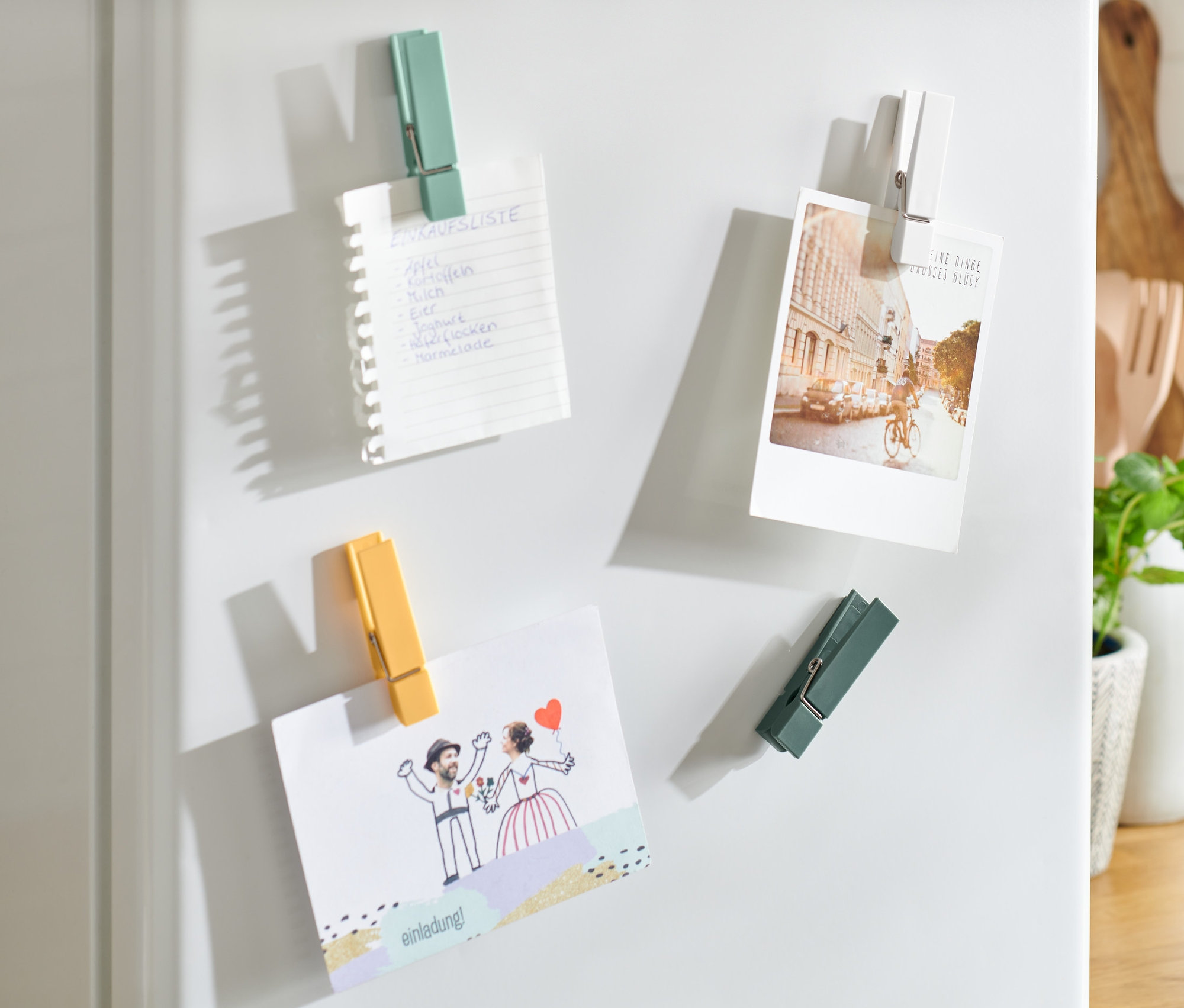 Na bielej chladničke sú pomocou 4 magnetov na chladničku pripevnené lístok s nákupným zoznamom, fotografia a svadobné oznámenie.