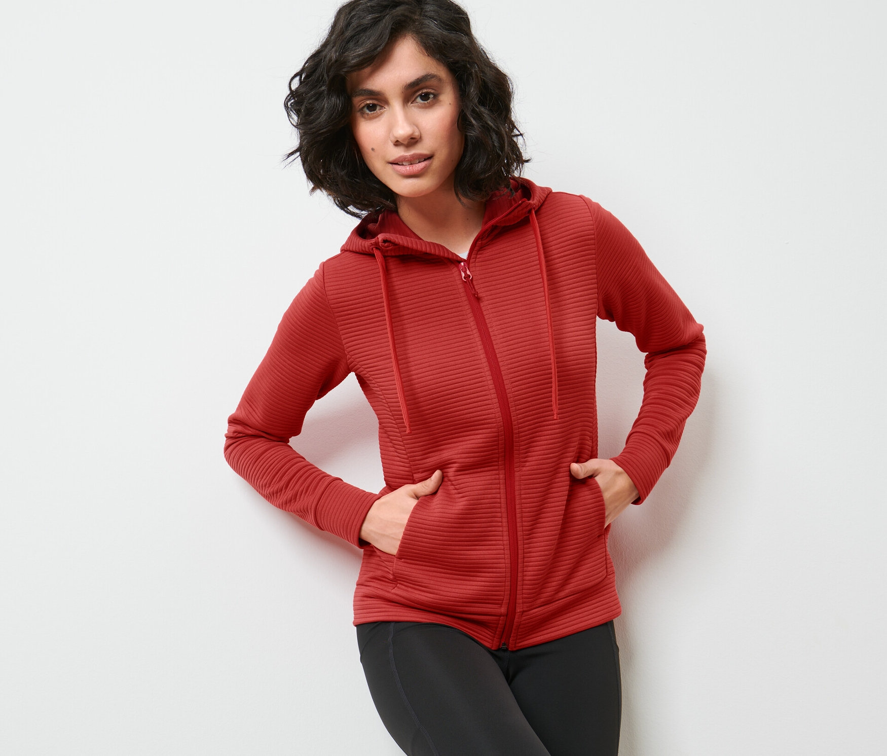 Žena pózuje v červenej mikine s kapucňou a čiernych legínach.