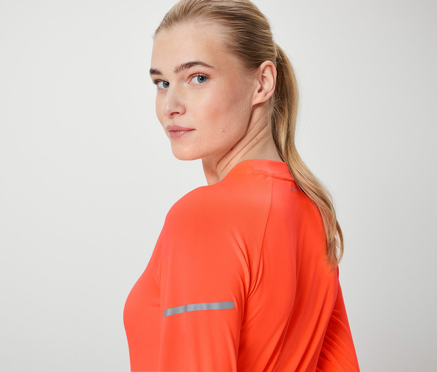 Blondína sa pozerá cez plece a má na sebe oranžový top s dlhým rukávom.