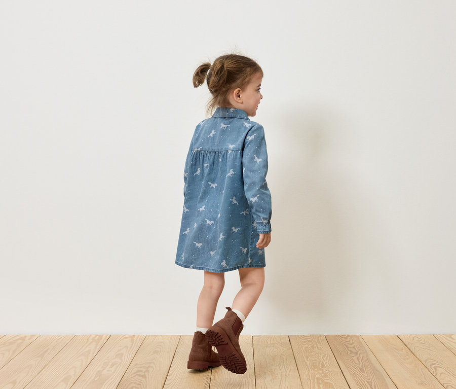 Dievča v džínsových šatách s potlačou koní a hnedých topánkach stojí bokom k bielej stene.