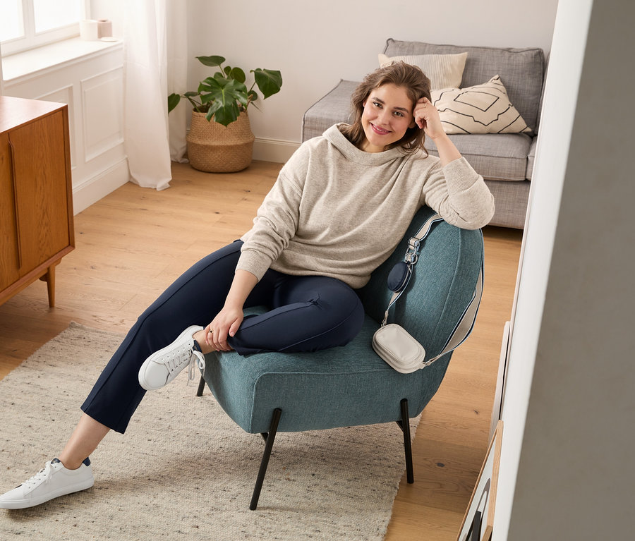 Žena sedí v kresle a má na sebe pulóver z jemnej pleteniny s kapucňou, legínsy s rozšírenými nohavicami a topánky sneaker z kože vypracovanej bez použitia chrómu. Cez kreslo je prehodená taška na nosenie krížom cez hrudník.