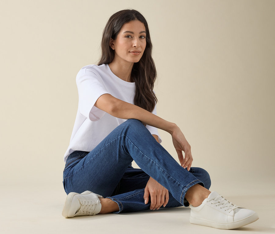 Žena sedí na zemi v slim džínsoch – strih »Emma«, modrý denim, biele tričko a tenisky.