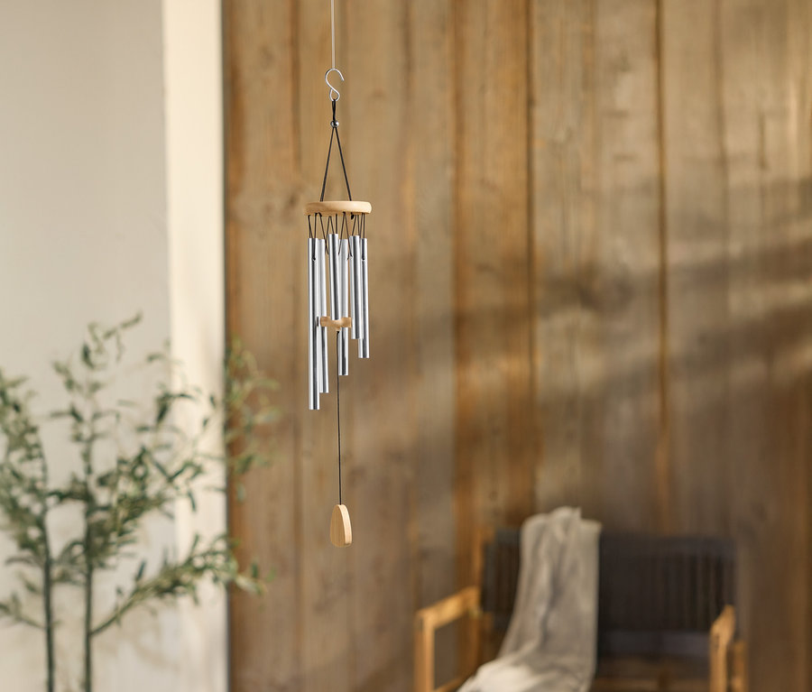 Zvonkohra so striebornými rúrkami visí pred drevenou stenou, vedľa rastliny a stoličky.