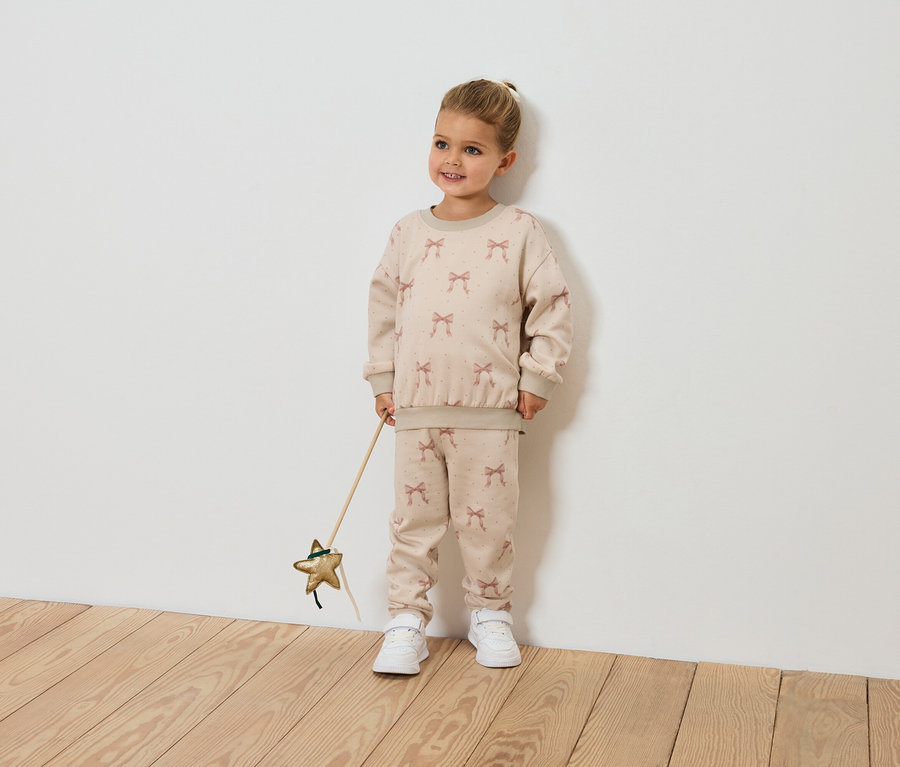 Usmievajúce sa dievčatko drží prútik v tvare hviezdy a má na sebe béžovú súpravu so vzorom mašlí.