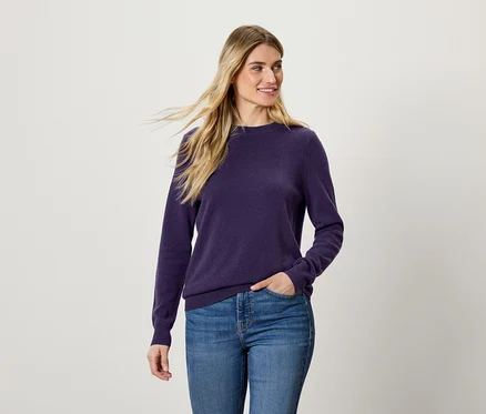 Žena s blond vlasmi má na sebe fialový sveter a modré džínsy.