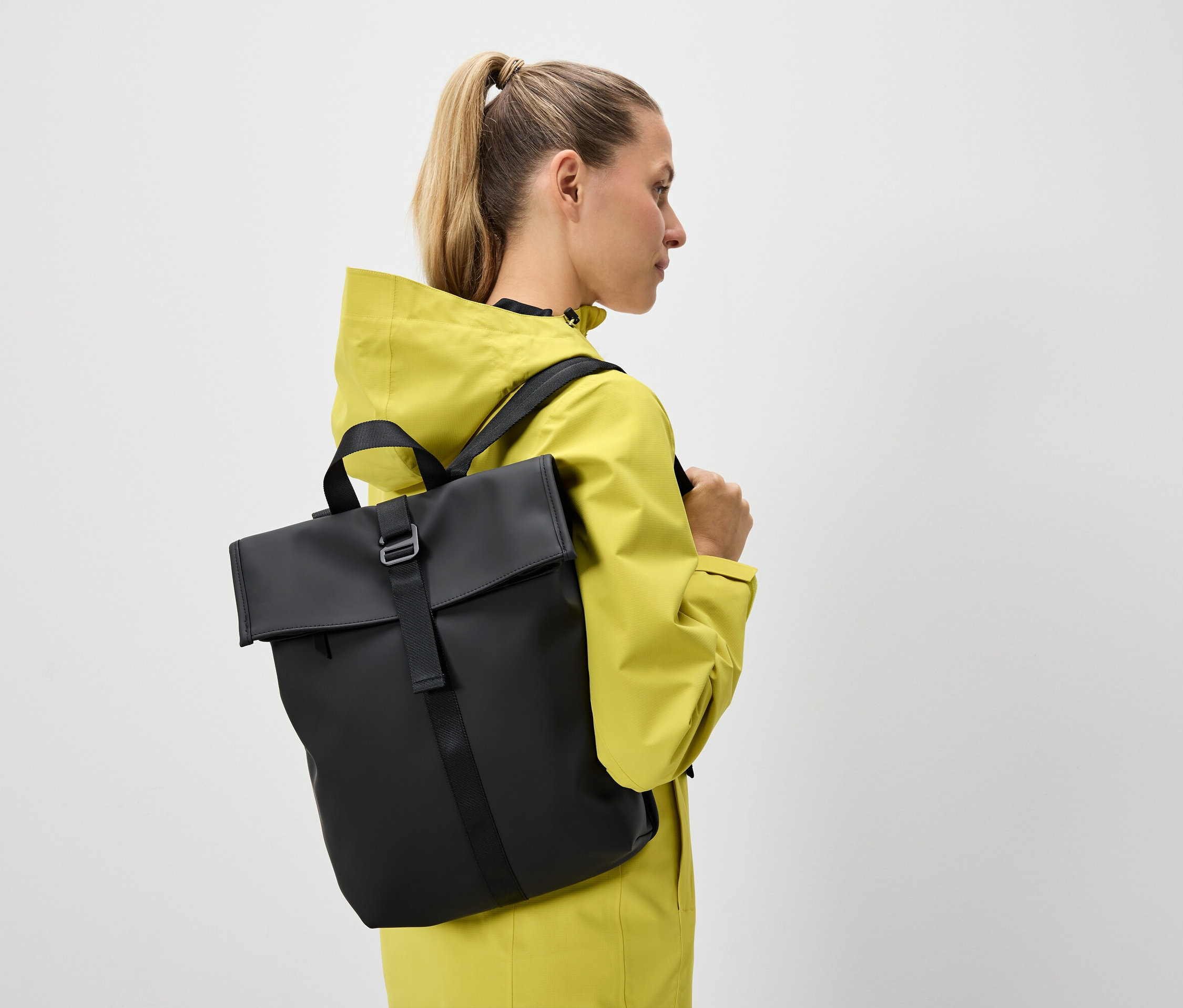 Žena s blond vlasmi stiahnutými do chvosta má na sebe žltú bundu a čierny batoh.