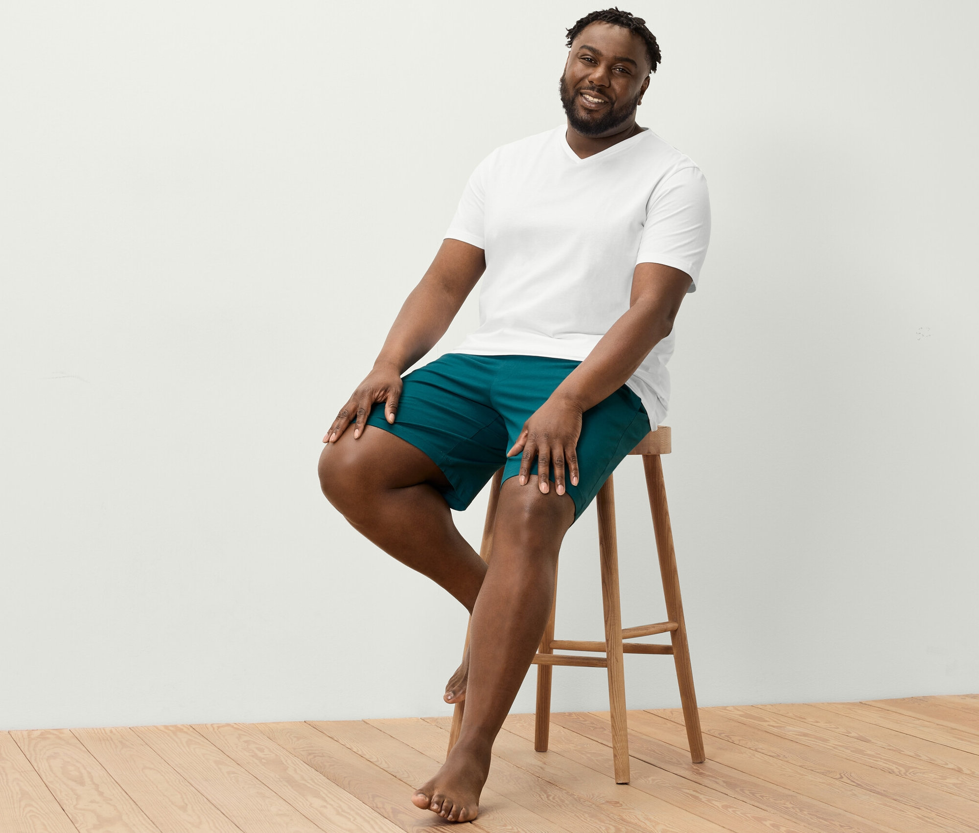 Muž sedí na stoličke a má na sebe biele tričko s výstrihom do V a pohodlné šortky.