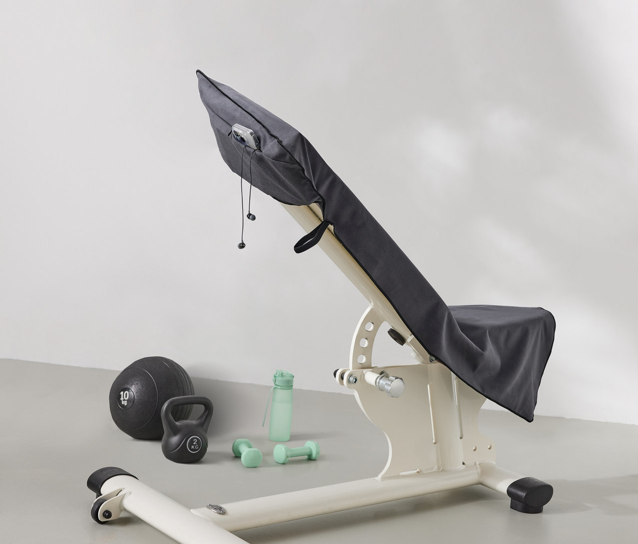 Posilňovacia lavica je zakrytá sivou látkou. Vedľa lavice leží medicinbal, kettlebell, fľaša na vodu a činky.