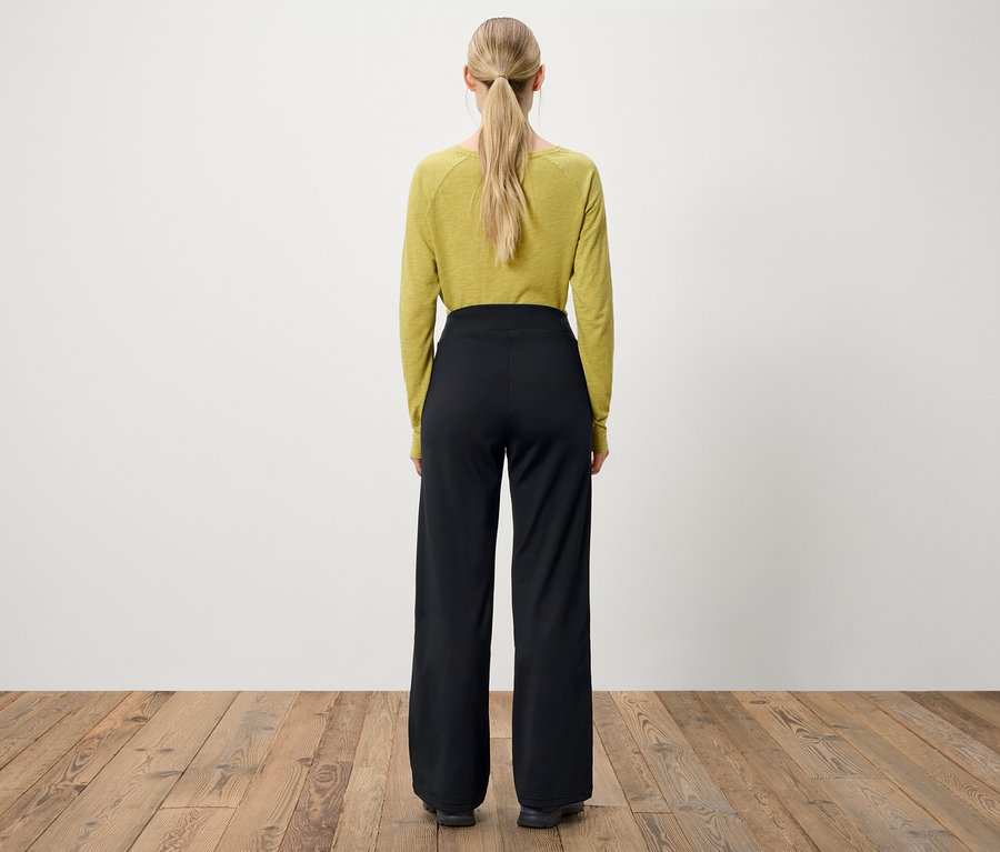 Zozadu je vidieť žena s blond vlasmi stiahnutými do copu, má na sebe olivovozelený top s dlhým rukávom a čierne nohavice so širokými nohavicami.