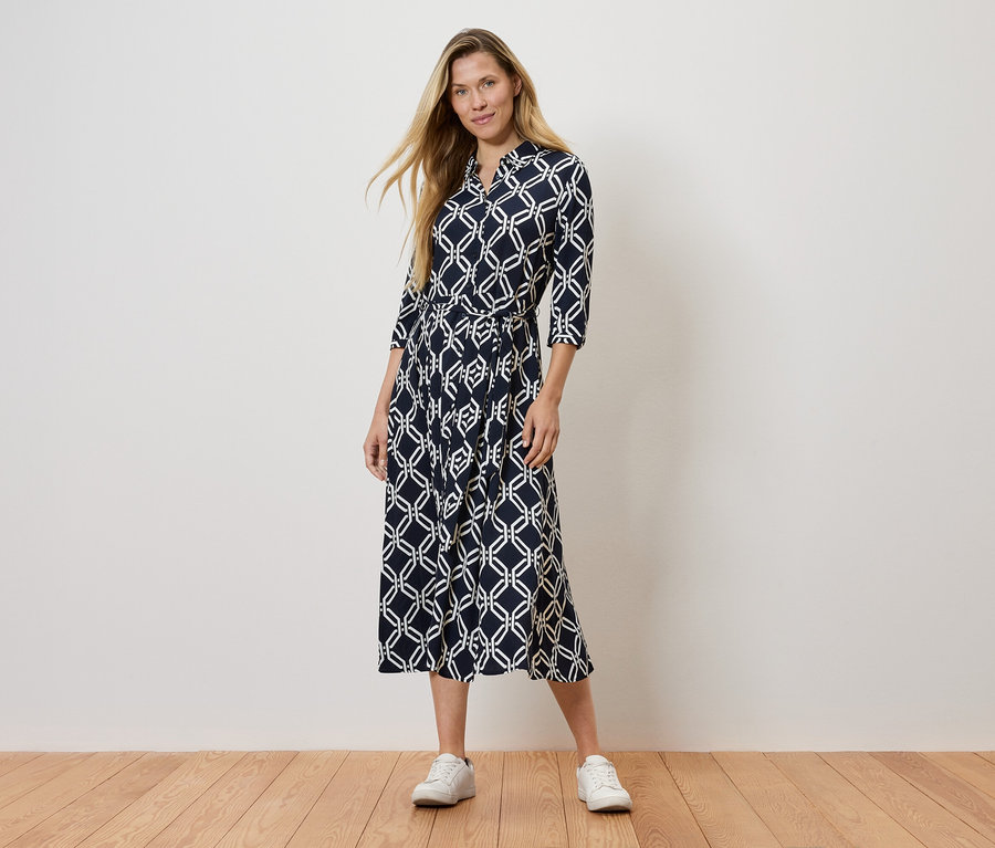 Žena stojí a má na sebe tmavomodré vzorované šaty a biele tenisky.