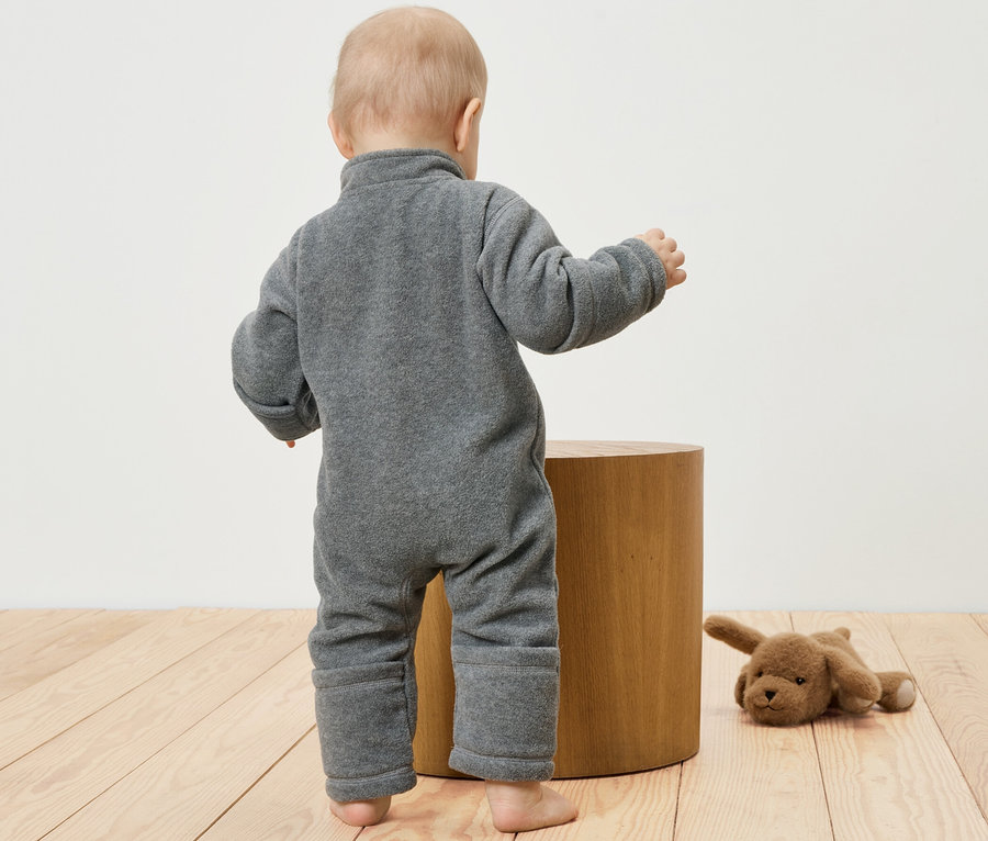 Batoľa v sivom flaušovom overale stojí pri drevenej stoličke. Vedľa leží plyšový pes.