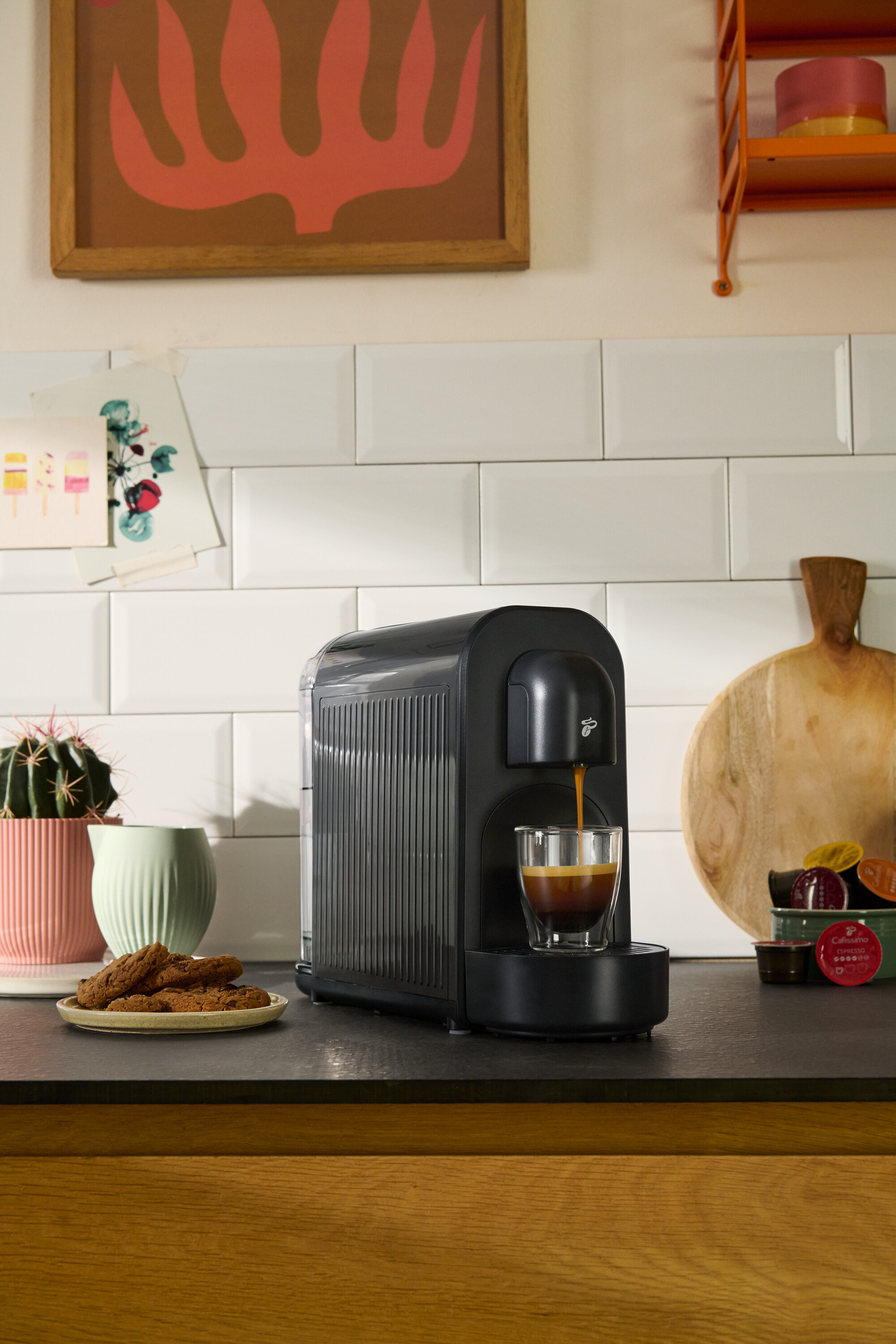 Čierny kávovar plní šálku kávou. Vidieť aj sušienky, sukulent a kuchynské dekorácie.