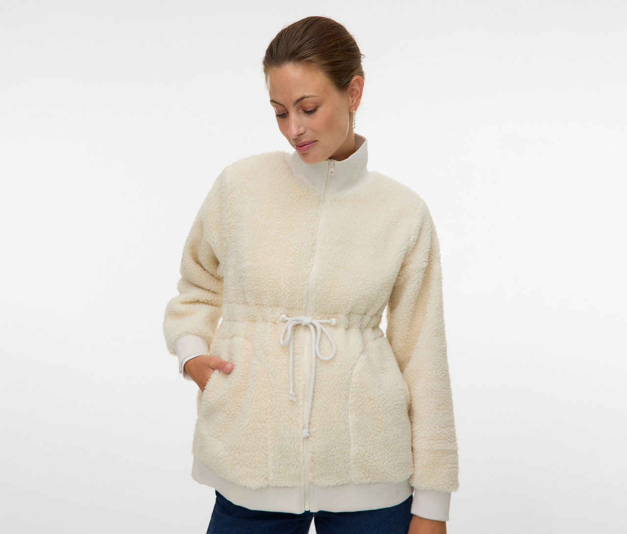 Žena má na sebe rozopnutú, krémovú plyšovú bundu MAMALICIOUS 3 v 1 »MLCARLY« so šnúrkou v páse a rukami vo vreckách.