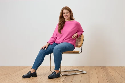Žena sedí na stoličke a má na sebe ružový sveter, modré džínsy a čierne mokasíny.