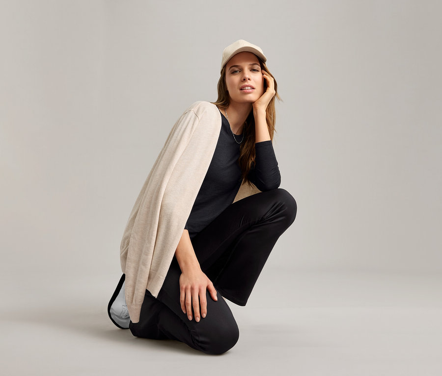 Žena sedí na podlahe v čiernom outdoorovom funkčnom tričku, béžovom kardigáne a svetlej šiltovke.