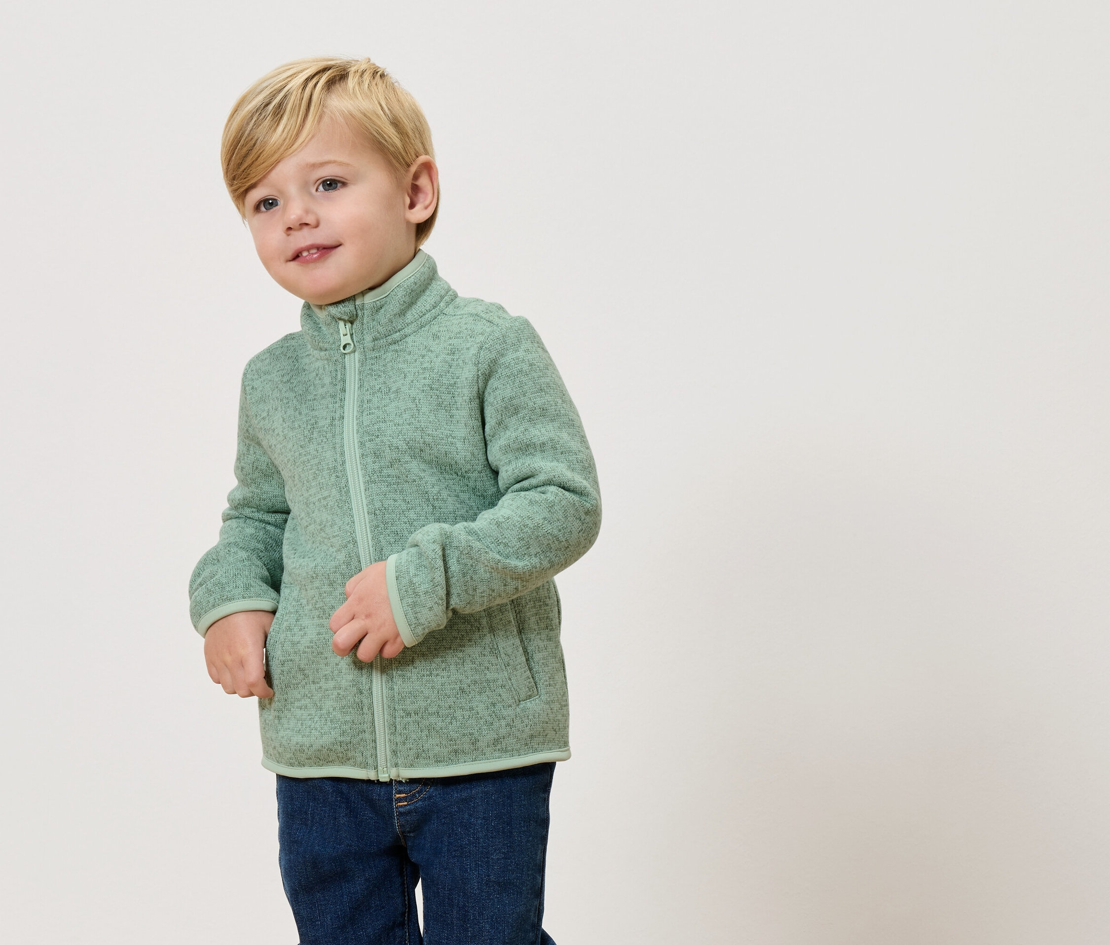 Blond chlapec má na sebe svetlozelenú flísovú bundu a džínsy.