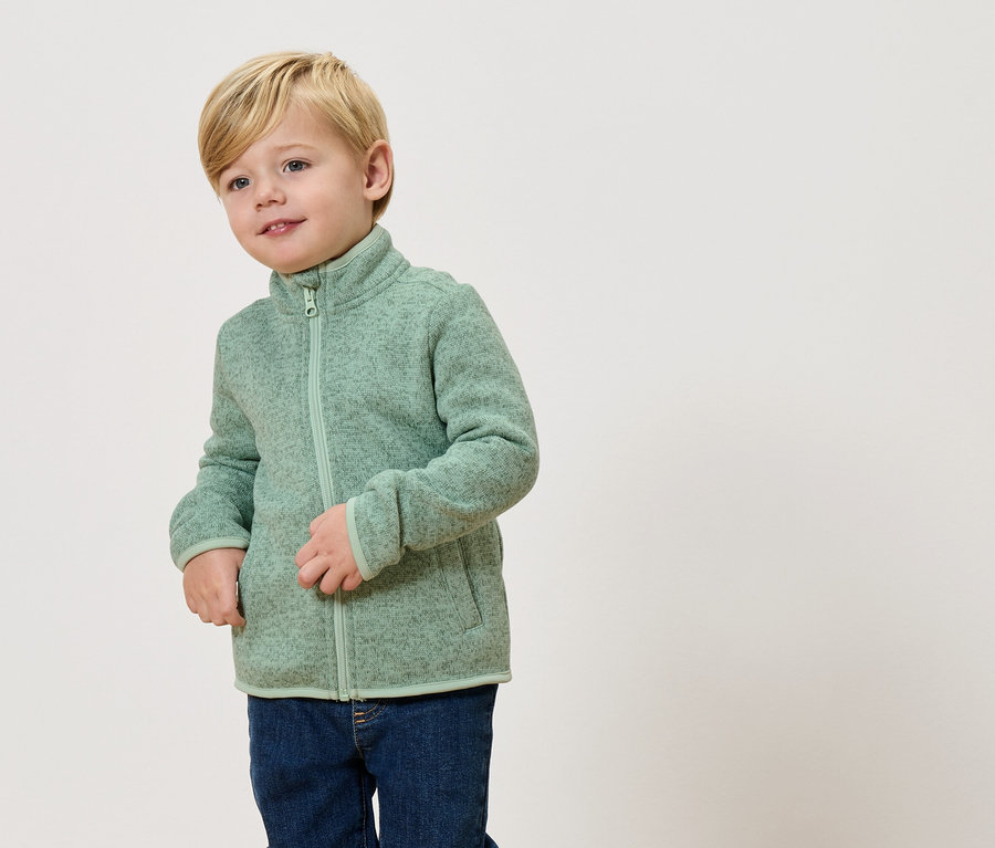 Blond chlapec má na sebe svetlozelenú flísovú bundu a džínsy.