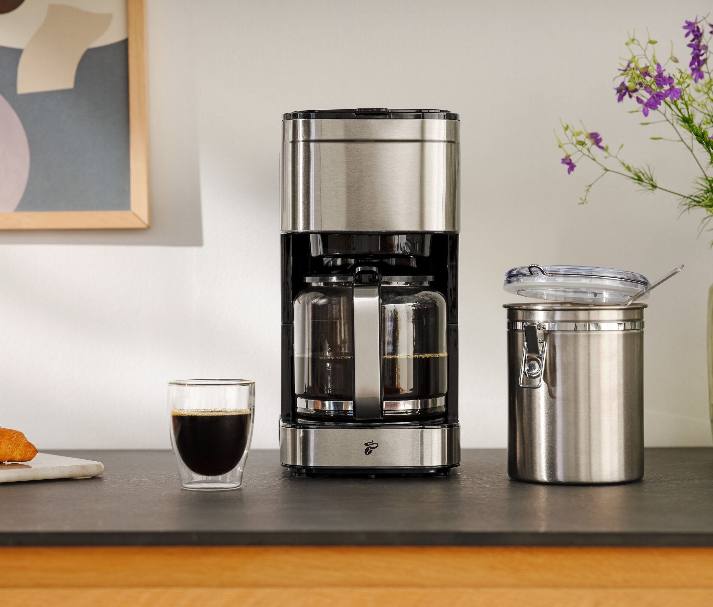 Kávovar vedľa pohára kávy, croissantu a nádoby na uskladnenie.