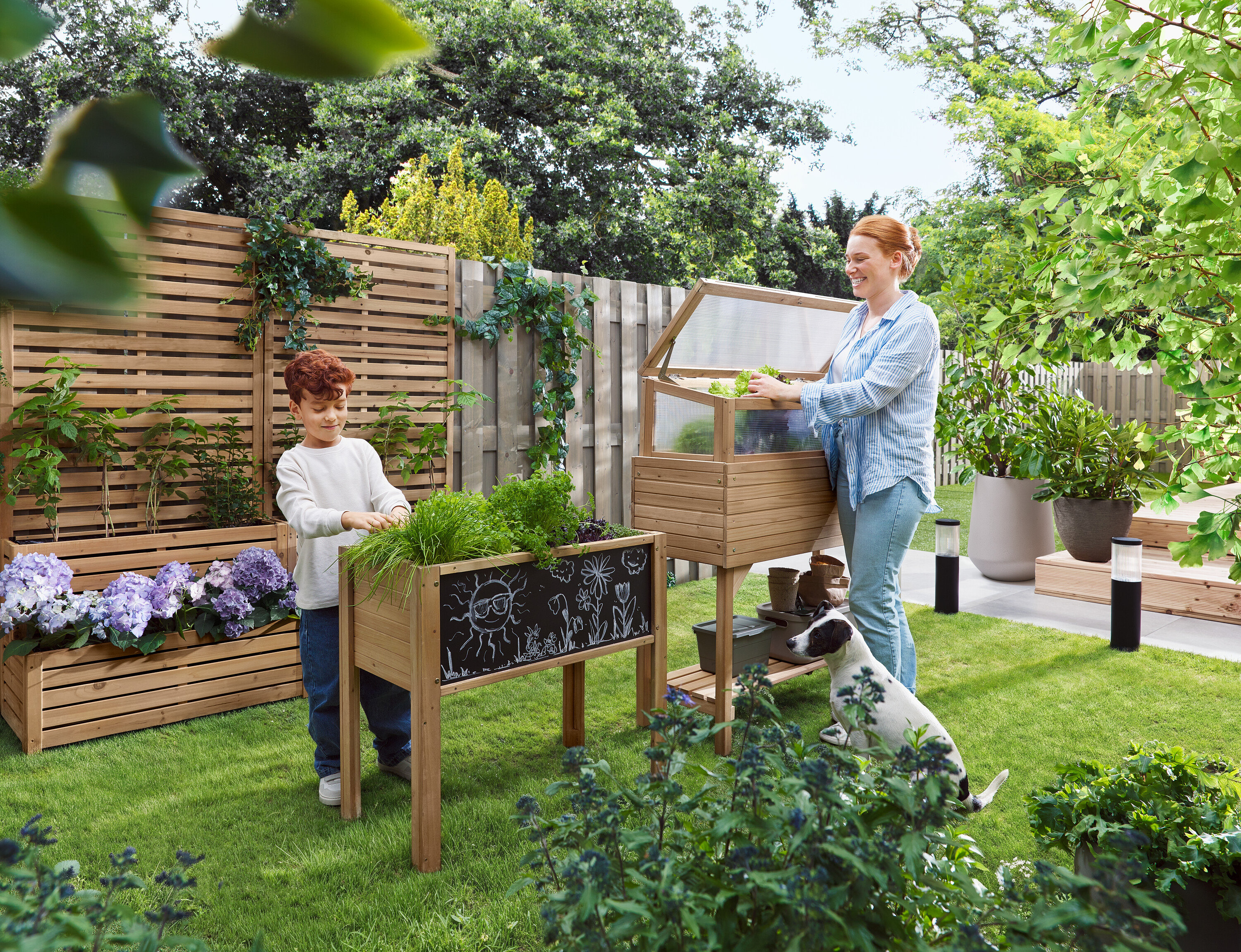 Žena otvára parenisko, zatiaľ čo dieťa stojí vedľa vyvýšeného záhonu. Pes sedí v tráve.