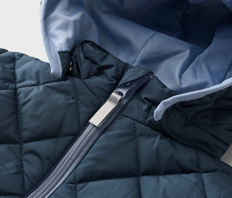 Detailný záber prešívanej, námorníckej modrej bundy so svetlomodrou podšívkou a strieborným zipsom.