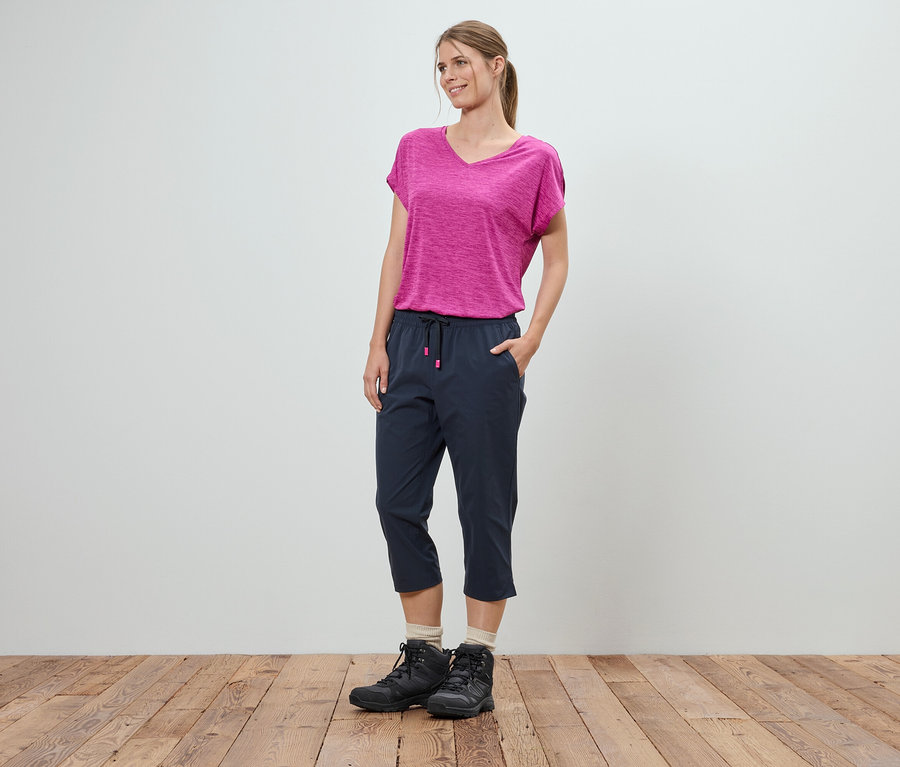 Žena stojí s rukami vo vreckách pred bielou stenou. Má na sebe ružové tričko, modré trojštvrťové nohavice a čierne trekové topánky.