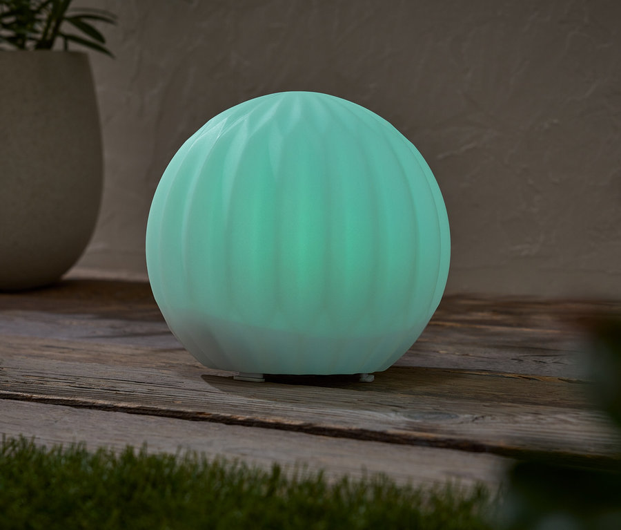 Svietiaca tyrkysová guľovitá lampa stojí na drevenom stole vedľa kvetináča s rastlinou.