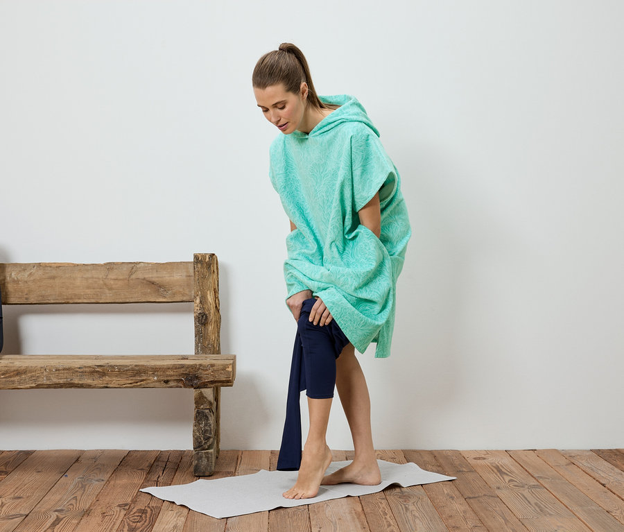 Žena sa prezlieka do nohavíc pod tyrkysovým pončom. V pozadí je lavička a batoh.