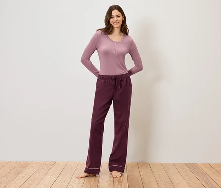 Žena v mauve tričku s dlhým rukávom a vínových nohaviciach so šnúrkou stojí na drevenej podlahe.