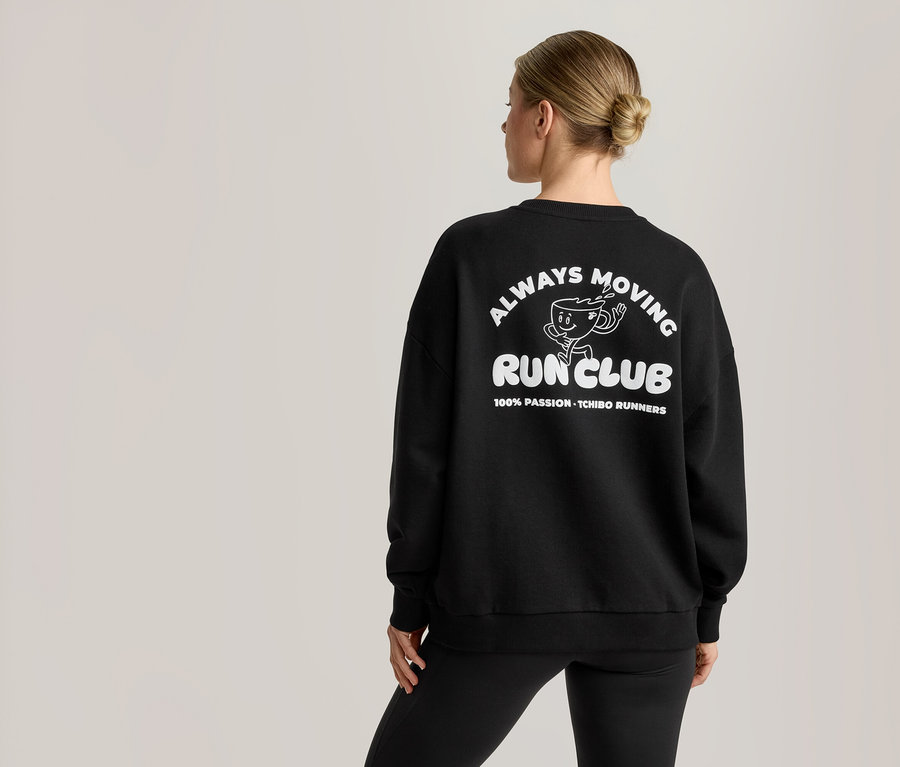 Žena videná zozadu má na sebe čiernu mikinu s bielou grafikou. Grafika obsahuje kreslenú postavičku šálky kávy a text 'ALWAYS MOVING RUN CLUB' a '100% PASSION - TCHIBO RUNNERS'.