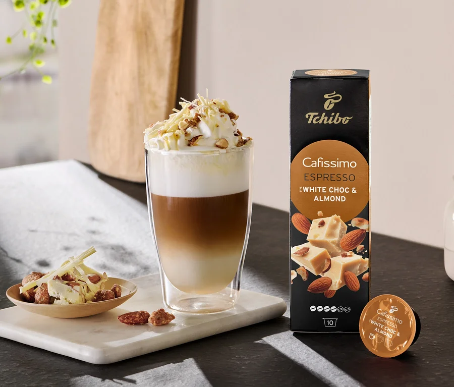 Vrstvené latte so šľahačkou a posýpkou vedľa balenia Cafissimo Flavoured Espresso - White Choc & Almond.
