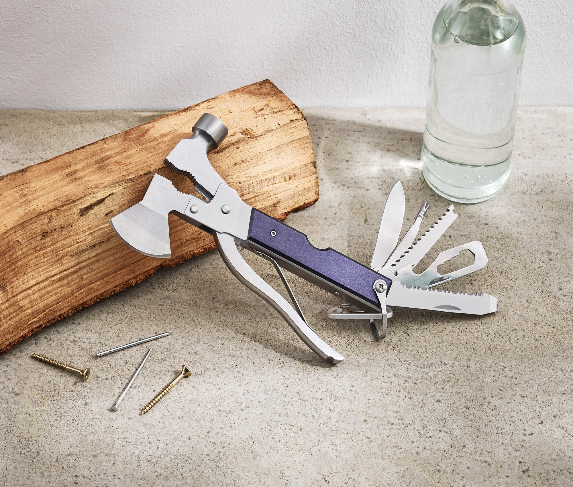 Na stole leží multifunkčný nástroj, kus dreva, klince, skrutky a fľaša s vodou.
