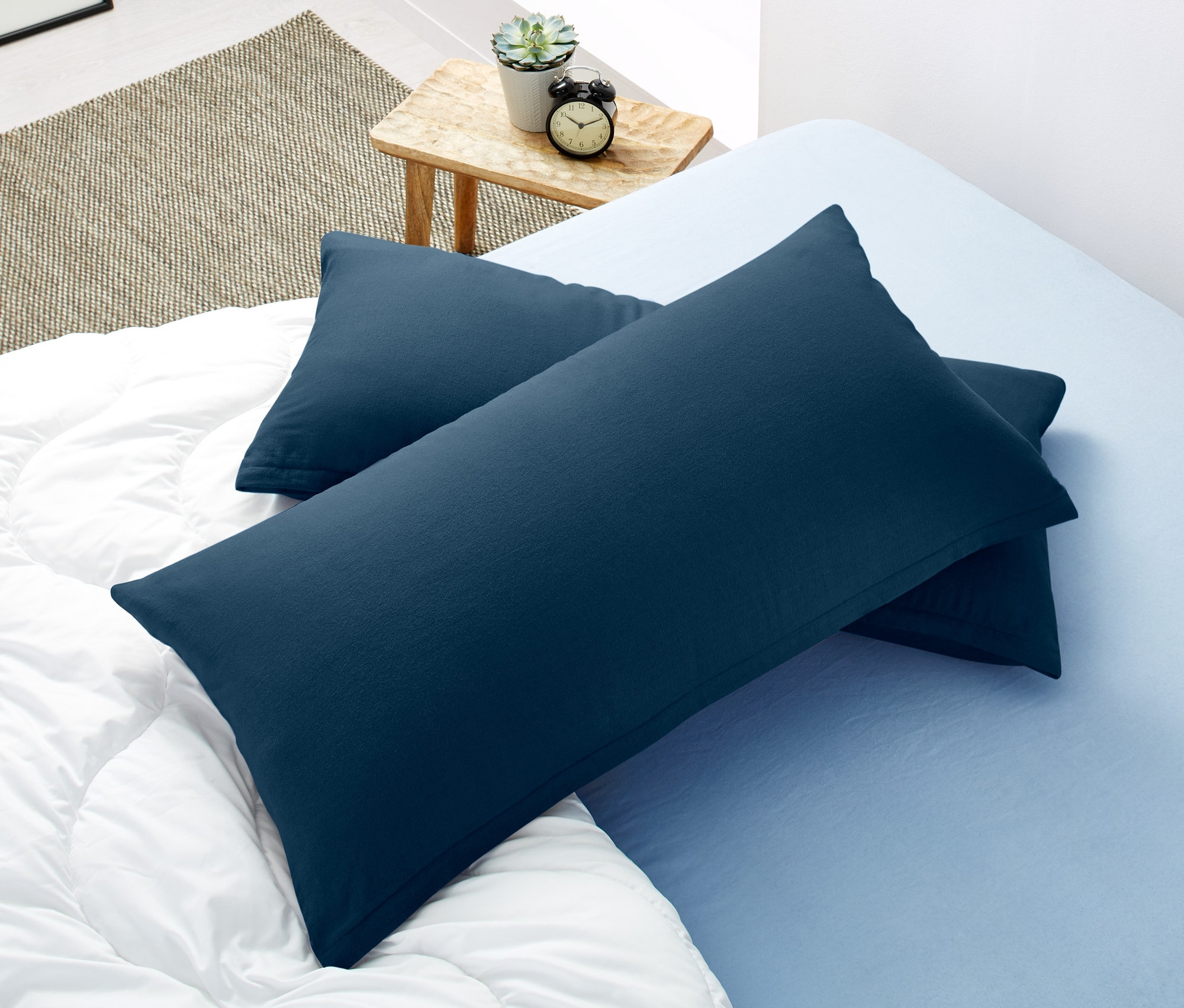 Dve tmavomodré obliečky na vankúše z česanej bavlny ležia na posteli s bielou prešívanou prikrývkou a svetlomodrou plachtou.