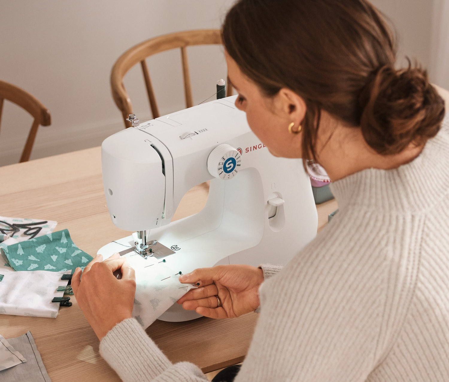 Žena šije látku na šijacom stroji SINGER vedľa súpravy nití, súpravy na šitie »Látkový adventný kalendár« a úložných boxov.