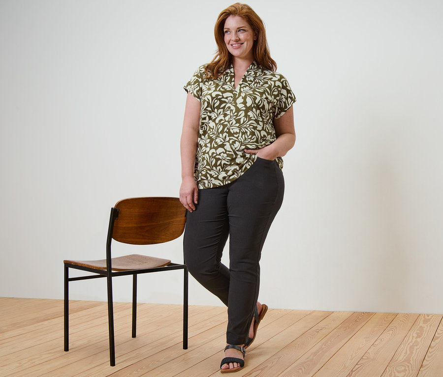 Žena stojí a pózuje v blúzkovom tričku s potlačou s výstrihom do V a v čiernych bengalínových nohaviciach, ruku má vo vrecku a vedľa nej stojí stolička.