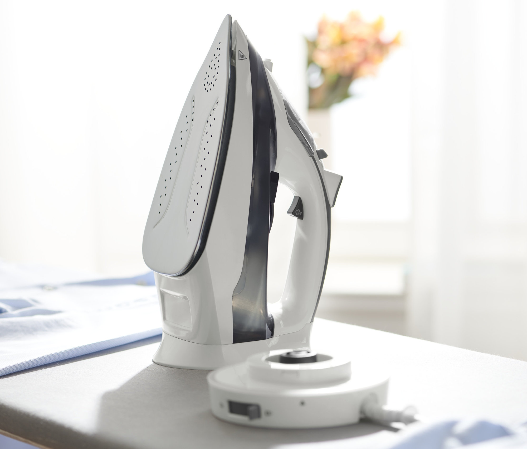Biela bezkáblová naparovacia žehlička stojí na sivej žehliacej doske vedľa kopy modrého oblečenia.