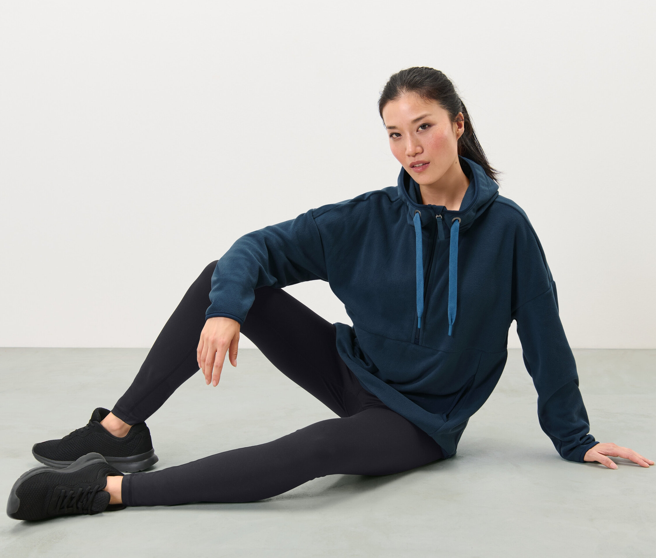 Žena sedí na zemi a má na sebe flaušový pulóver s kapucňou, čierne legíny a tenisky.