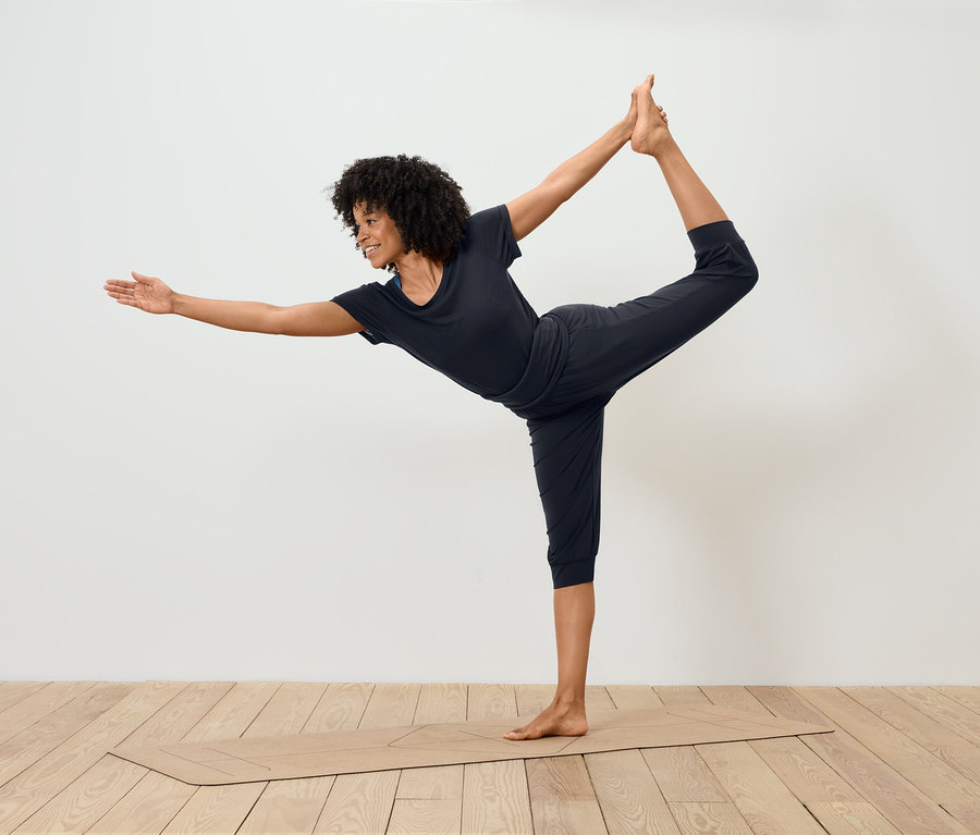 Žena v čiernom športovom oblečení stojí na podložke na jogu v pozícii tanečnice.