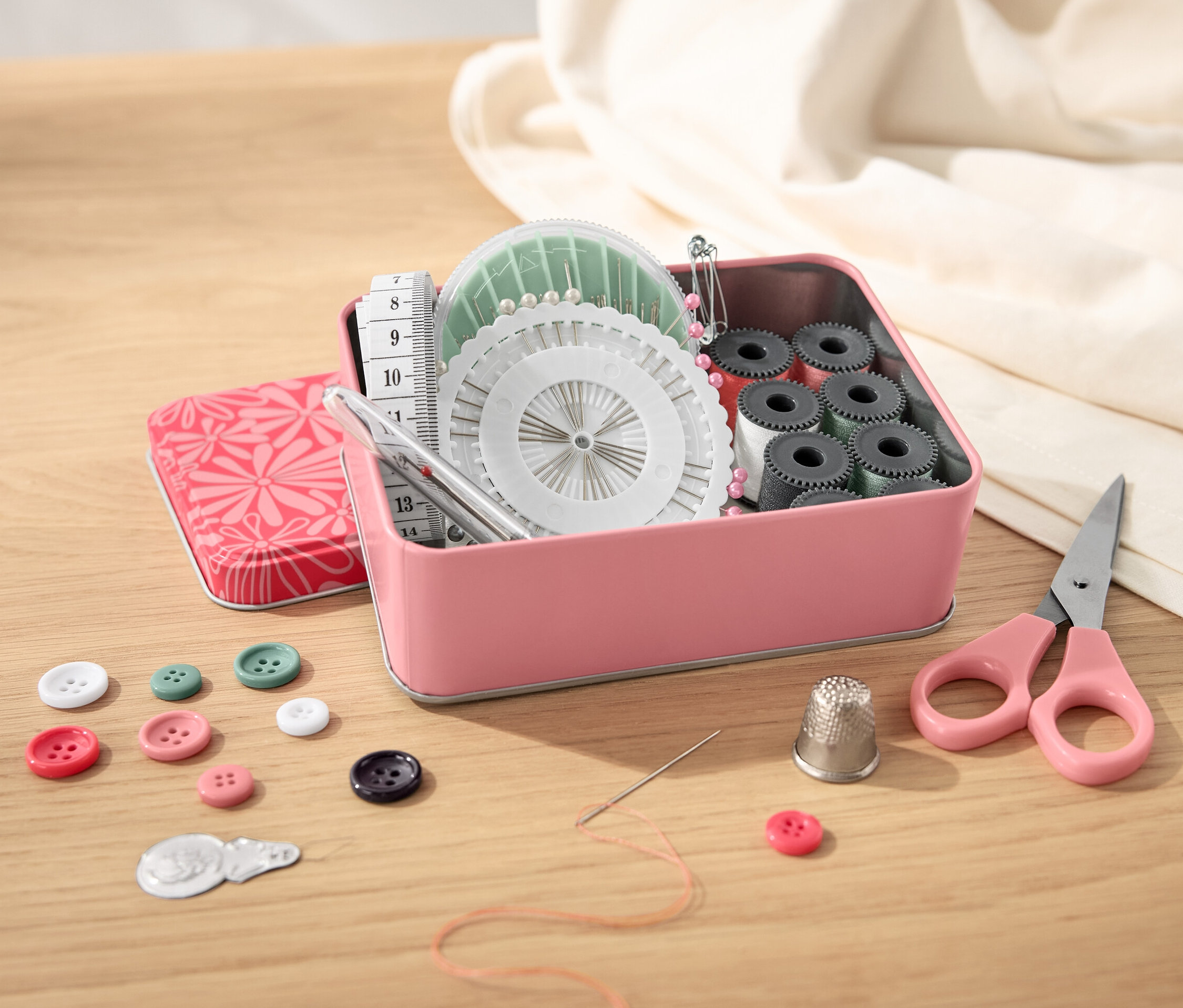Otvorená kovová krabička so šijacou súpravou, gombíkmi, nožnicami a krajčírskym metrom na drevenom stole.