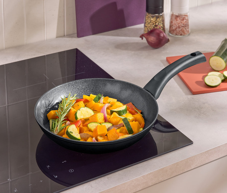 Panvica s restovanou zeleninou na indukčnej varnej doske. Vedľa panvice leží na doske nakrájaná cuketa.