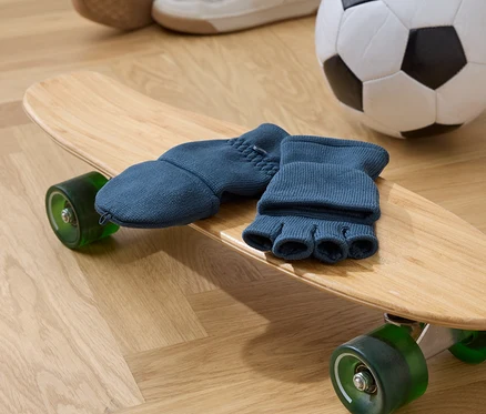 Modré prstové rukavice ležia na drevenom skateboarde so zelenými kolieskami. V pozadí je futbalová lopta a topánky.
