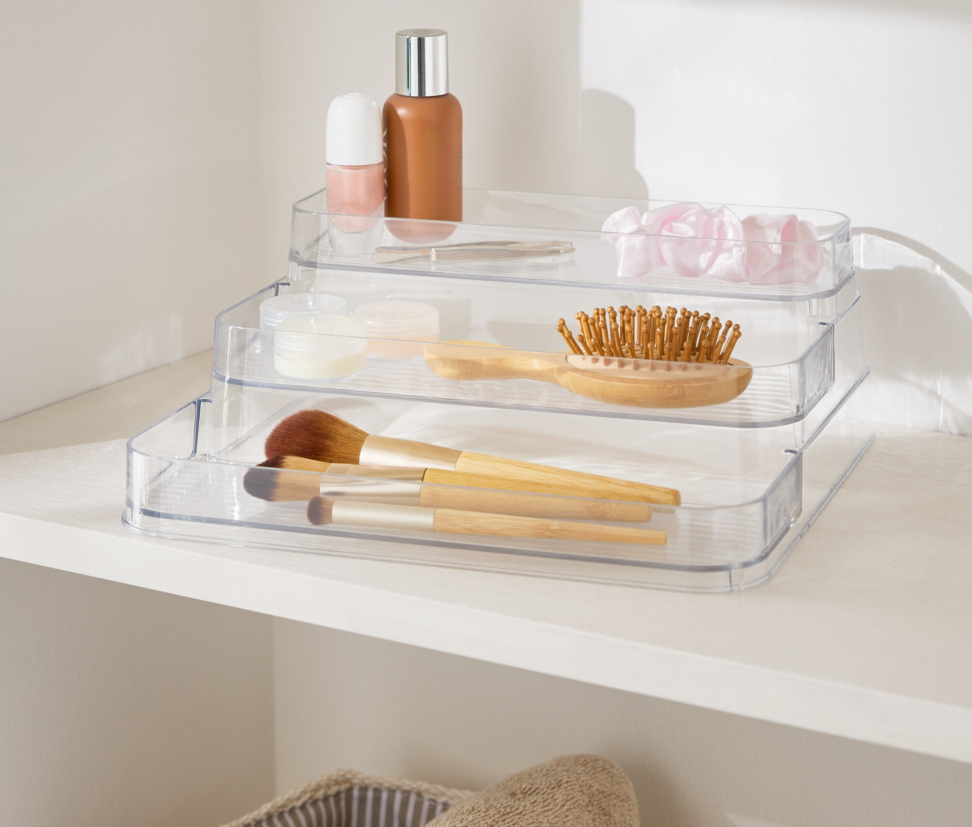 Trojposchodový priehľadný organizér na líčidlá na bielej polici vedľa košíkov s uterákmi.